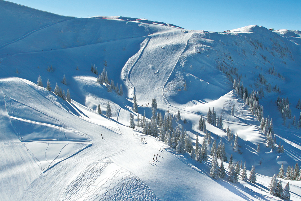 Aerial view of a snowy mountain landscape with ski tracks and trails. Several skiers and snowboarders can be seen gliding down the slopes. Pine trees dot the area, and the sky is clear and blue, indicating a sunny winter day.