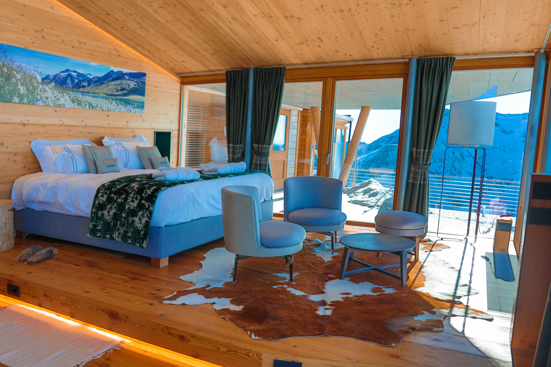 A cozy mountain lodge room with a large bed, wooden walls, and a cowhide rug. It features a sitting area with chairs and a stunning view of snowy mountains through glass doors. Decor includes a landscape picture above the bed.