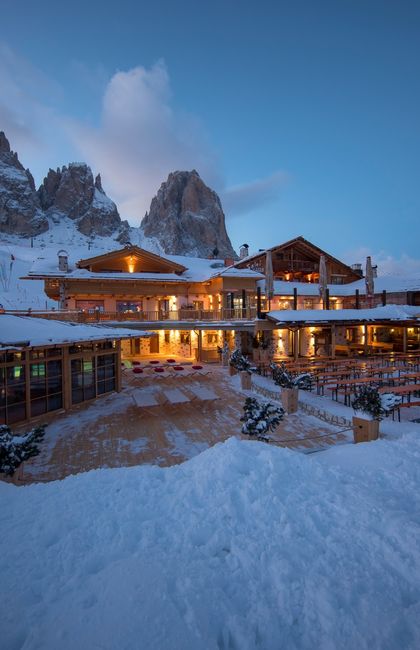 A snow-covered mountain lodge with warm lights glows against a backdrop of towering rocky peaks. The sky is a soft blue dusk, enhancing the cozy ambiance of the wooden structure. Outdoor seating is partially visible in the foreground.