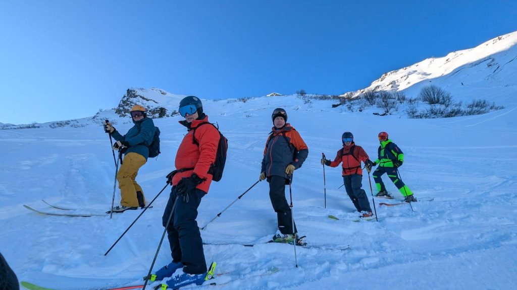 Four skiers in colorful winter gear stand in a row on a snowy mountain slope, holding ski poles, with clear blue sky and snow-covered peaks in the background.