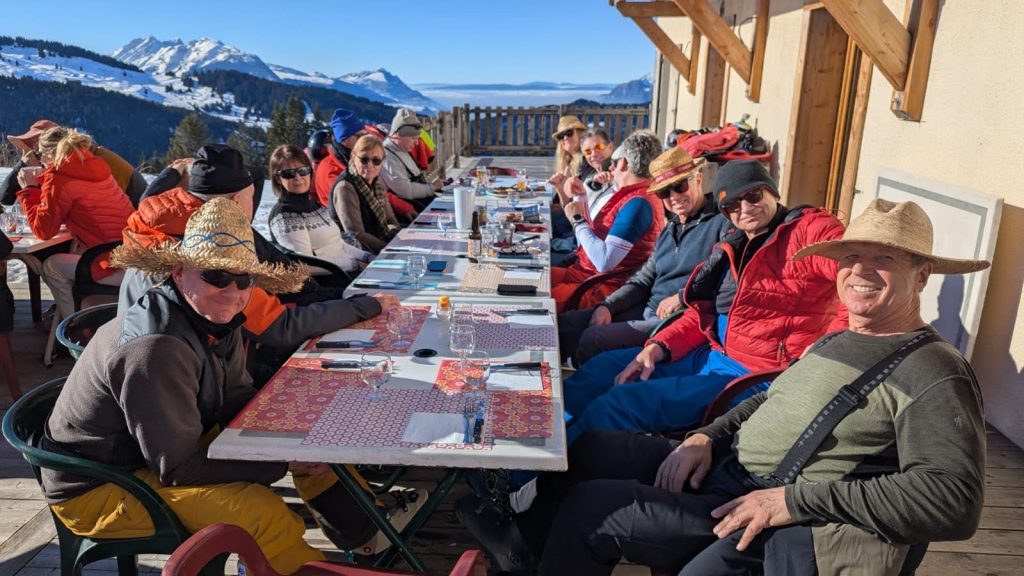 A group of people wearing hats sit around long tables outdoors, enjoying a sunny meal on a mountain terrace with snowy peaks and blue sky in the background.