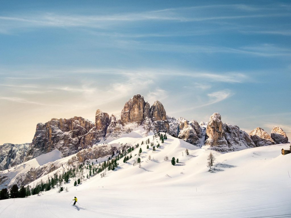 A person skis down a snowy slope dotted with small trees, with rugged mountain peaks in the background under a blue sky with wispy clouds.