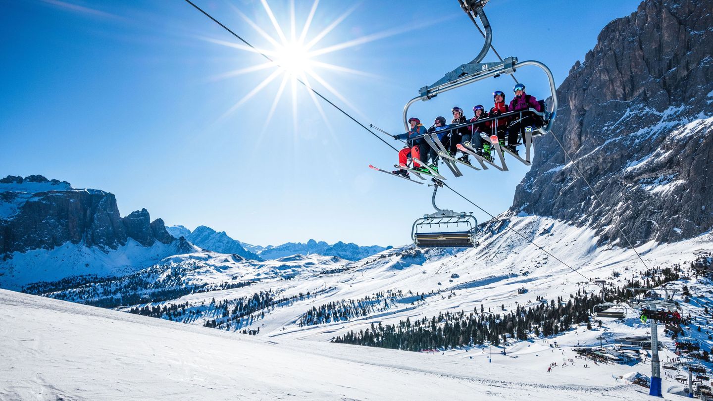 A group of skiers sits on a chairlift ascending a snowy mountain slope under a bright sun, with clear blue skies and rugged, snow-covered peaks in the background.